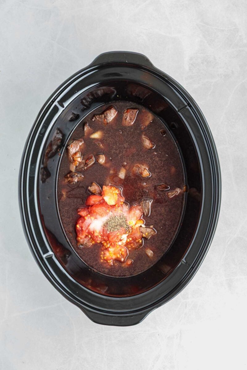 A slow cooker filled with chunks of meat, diced onions, tomato sauce, and seasonings, ready to be cooked.