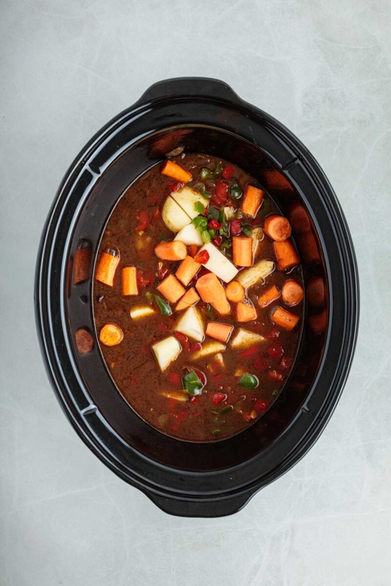 A slow cooker filled with broth, chopped carrots, potatoes, and diced tomatoes on a light surface.