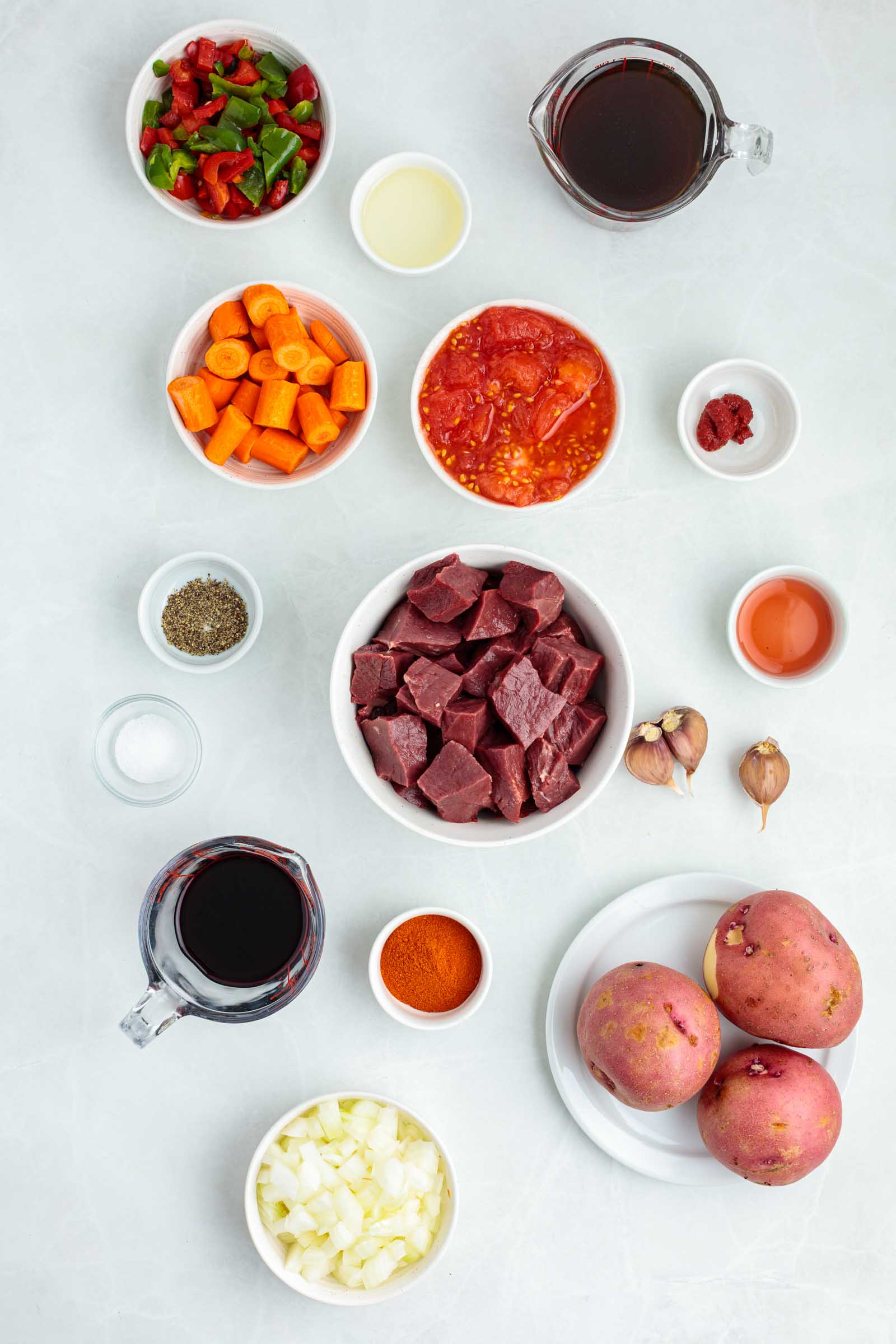 Various ingredients for a stew are arranged on a white surface, including cubed beef heart, potatoes, onion, carrots, garlic, spices, tomato, chopped peppers, and liquids in small bowls.