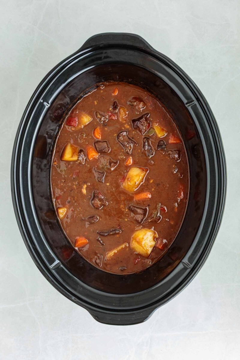 A slow cooker filled with beef stew containing chunks of beef, potatoes, carrots, and a dark brown broth.