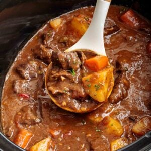 A wooden ladle scoops beef heart stew with potatoes and carrots from a slow cooker, showing chunks of meat and vegetables in a thick brown broth.