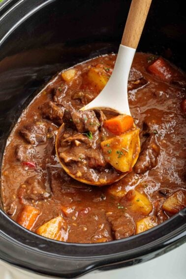A wooden ladle scoops beef heart stew with potatoes and carrots from a slow cooker, showing chunks of meat and vegetables in a thick brown broth.