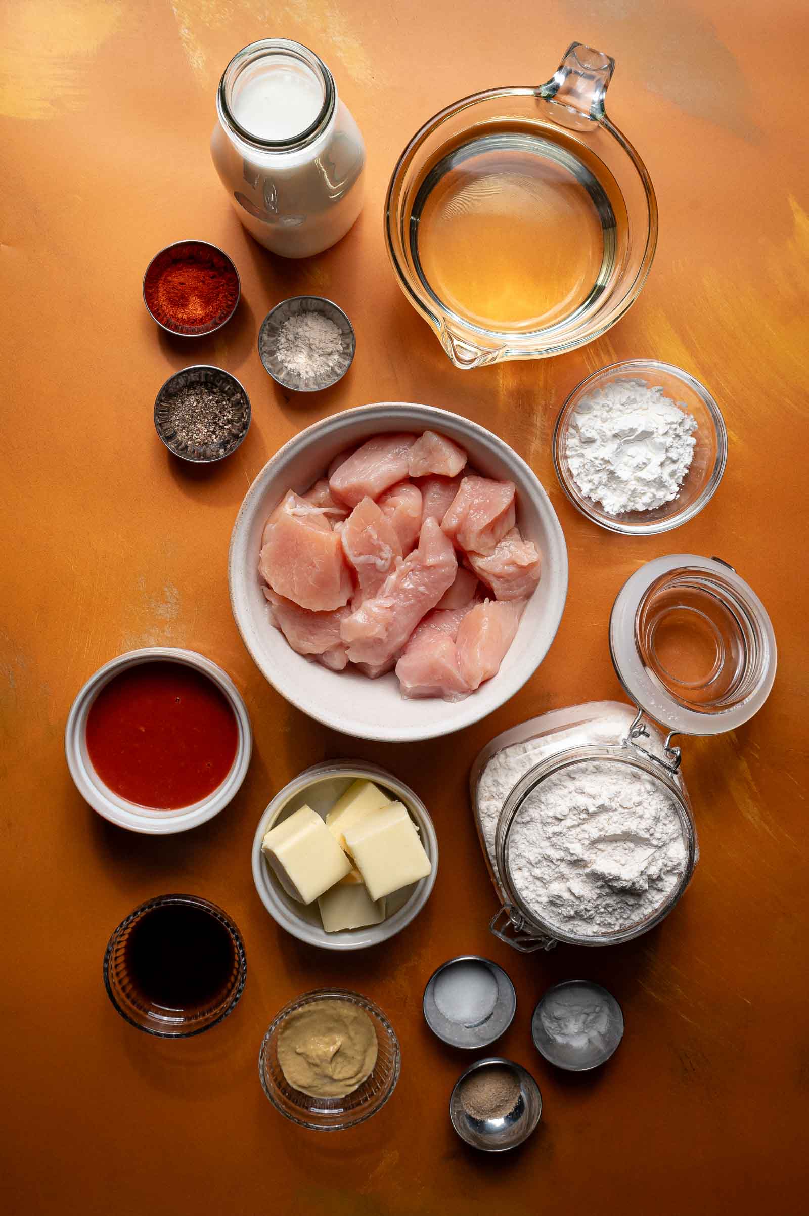 Raw cubed chicken, flour, butter, spices, sauces, milk, water, and other ingredients arranged on an orange surface for cooking preparation.