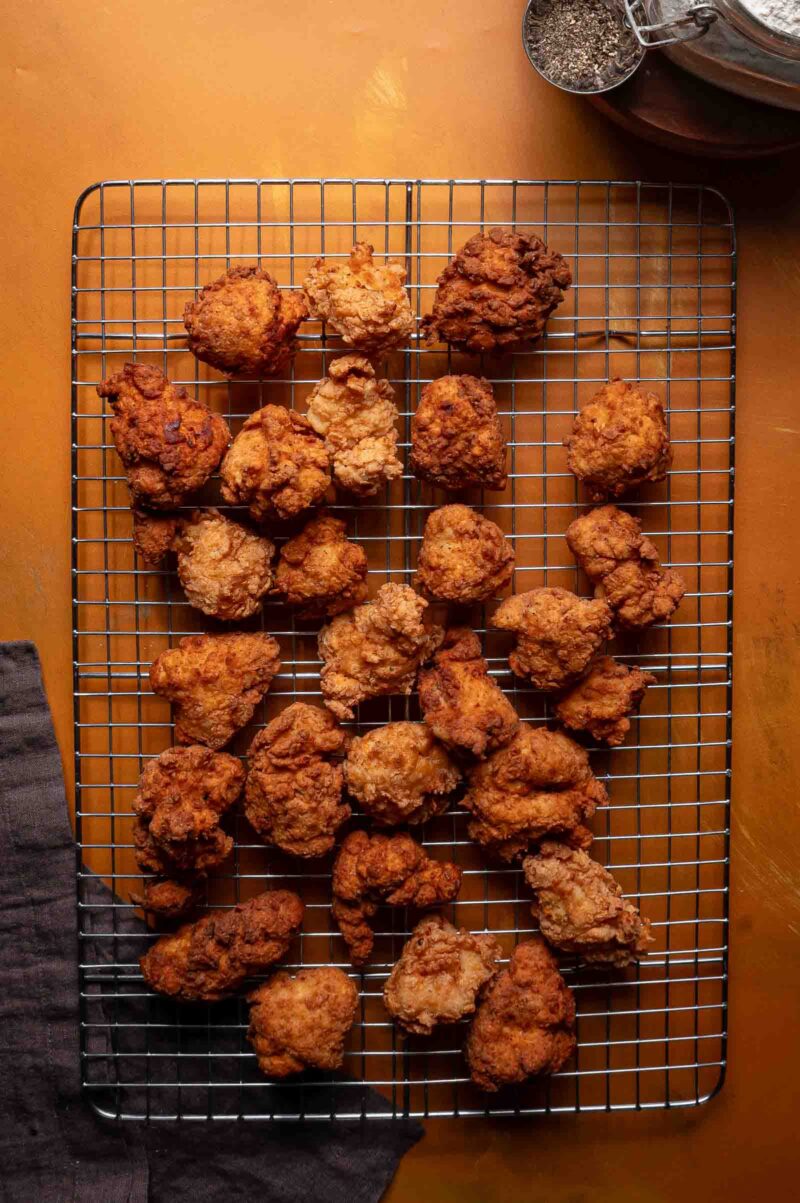 A cooling rack with several pieces of fried chicken placed on it, set on an orange surface with a black napkin on the left.