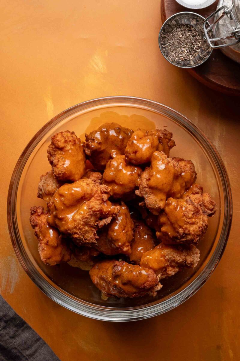 A glass bowl filled with fried chicken pieces coated in a buffalo sauce, placed on an orange surface with a small bowl of pepper nearby.