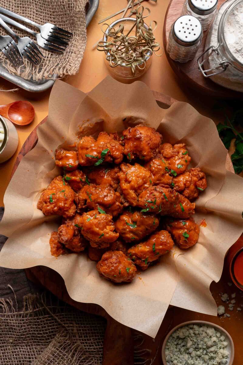 A basket lined with parchment paper filled with saucy boneless buffalo wings, garnished with chopped herbs, surrounded by various kitchen items and ingredients.