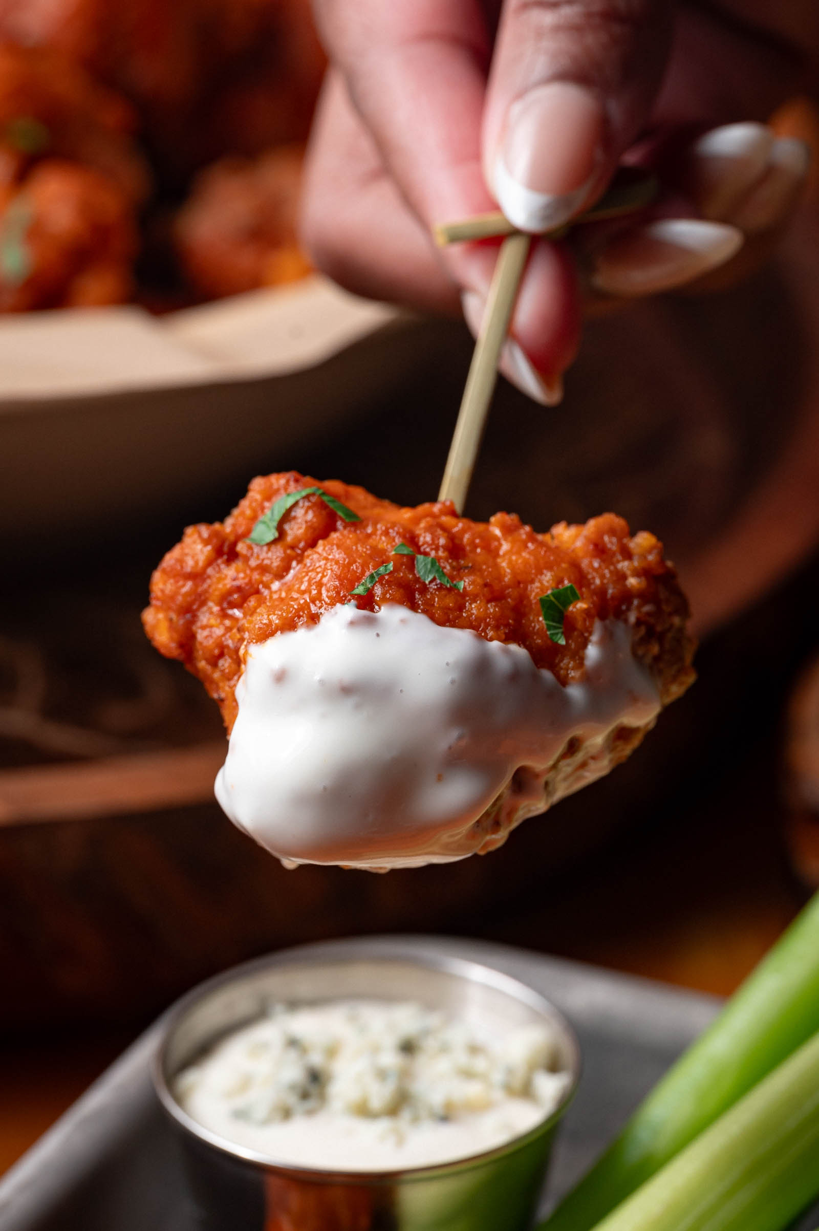 A hand holds a boneless buffalo chicken wing on a skewer, dipped in blue cheese sauce, with more wings and celery visible in the background.