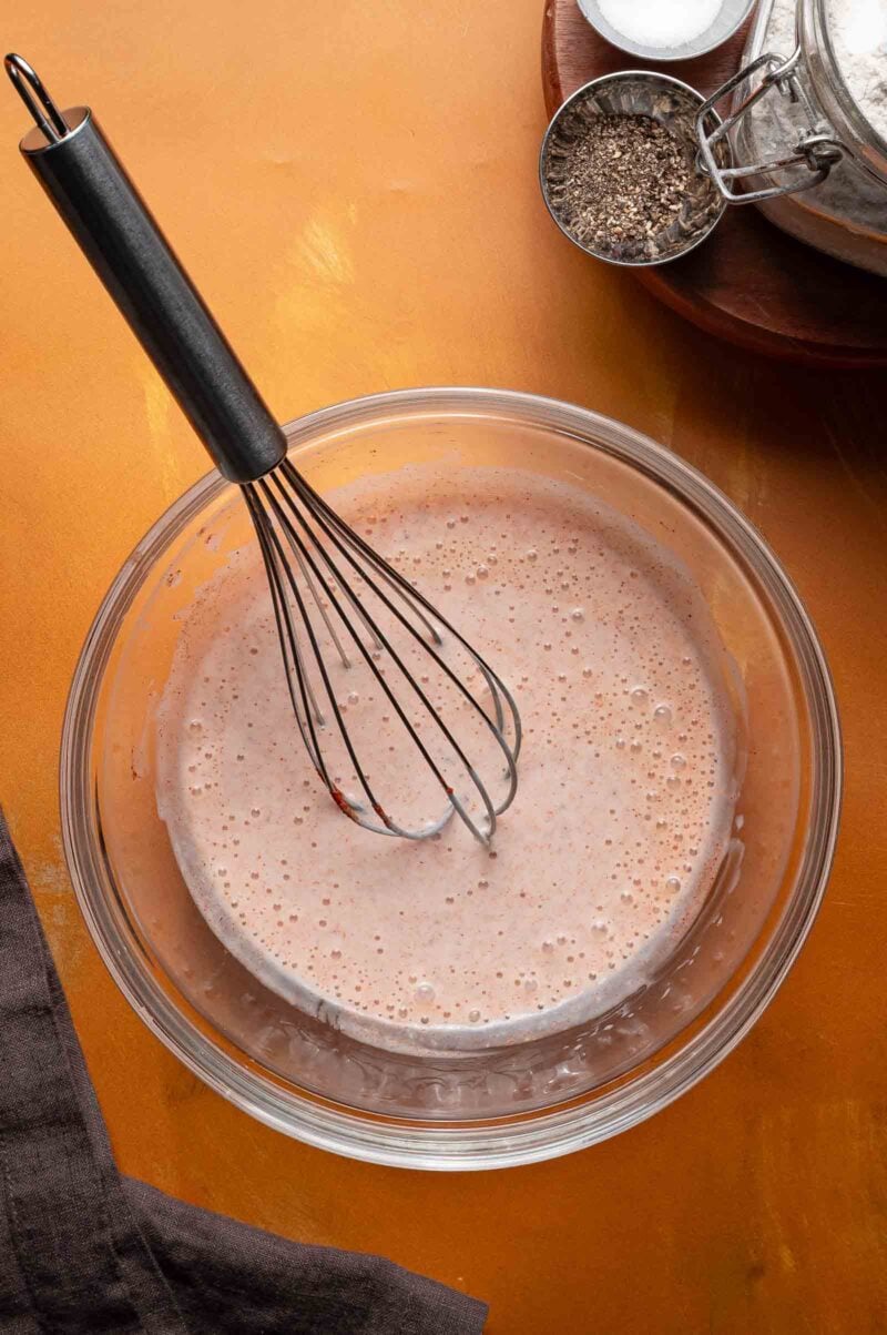 A glass bowl filled with a seasoned buttermilk chicken brine being mixed with a metal whisk, set on a yellow surface with some seasoning and a jar nearby.