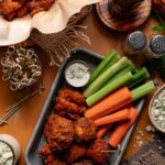 A tray of boneless buffalo chicken wings with blue cheese dip, celery, and carrot sticks; additional wings, a drink, and condiments are arranged around the table.