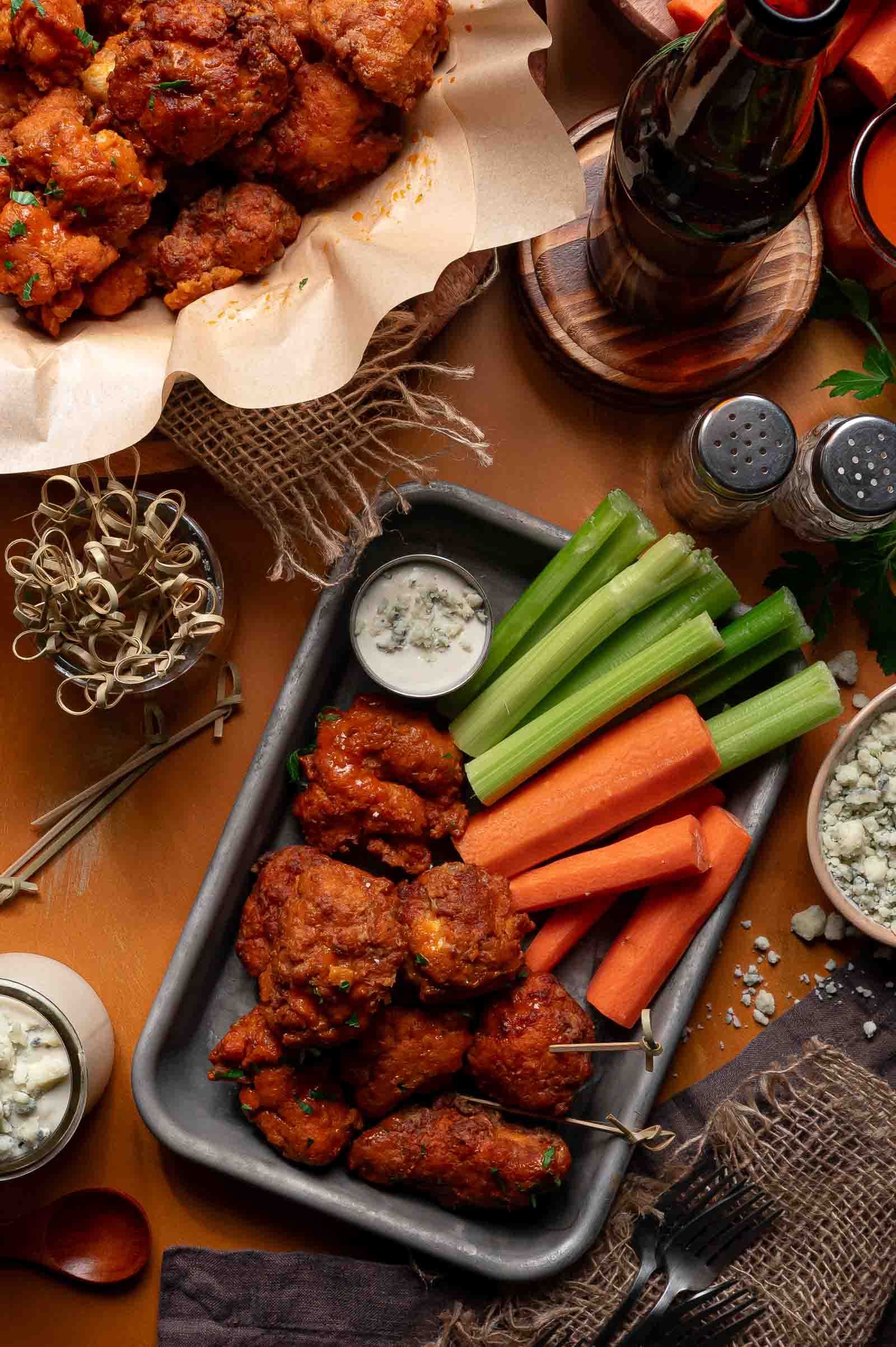 A tray of boneless buffalo chicken wings with blue cheese dip, celery, and carrot sticks; additional wings, a drink, and condiments are arranged around the table.