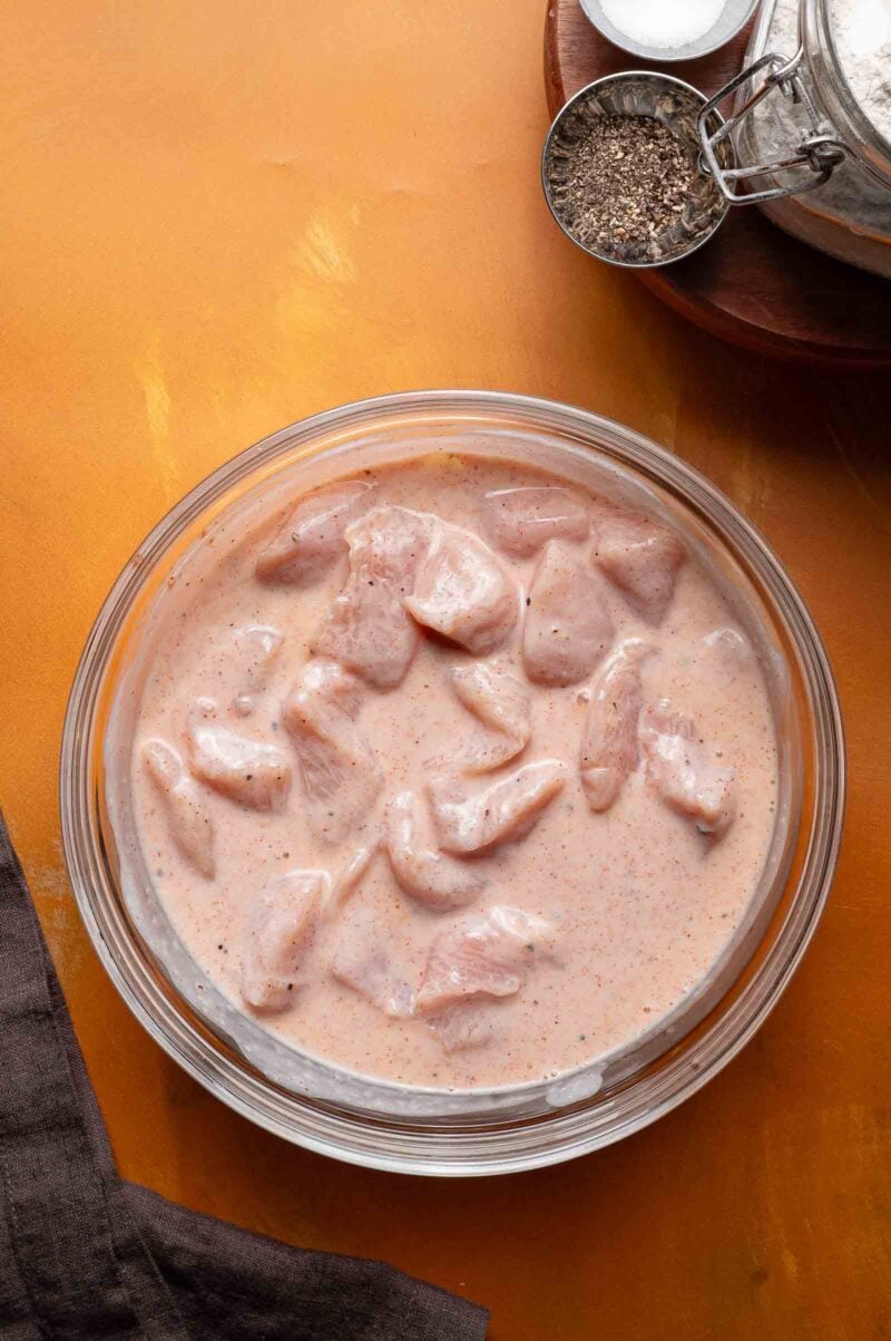 Raw chicken pieces marinating in a creamy, spiced buttermilk brine inside a clear glass bowl on a yellow surface. Seasonings are visible in a metal measuring cup nearby.