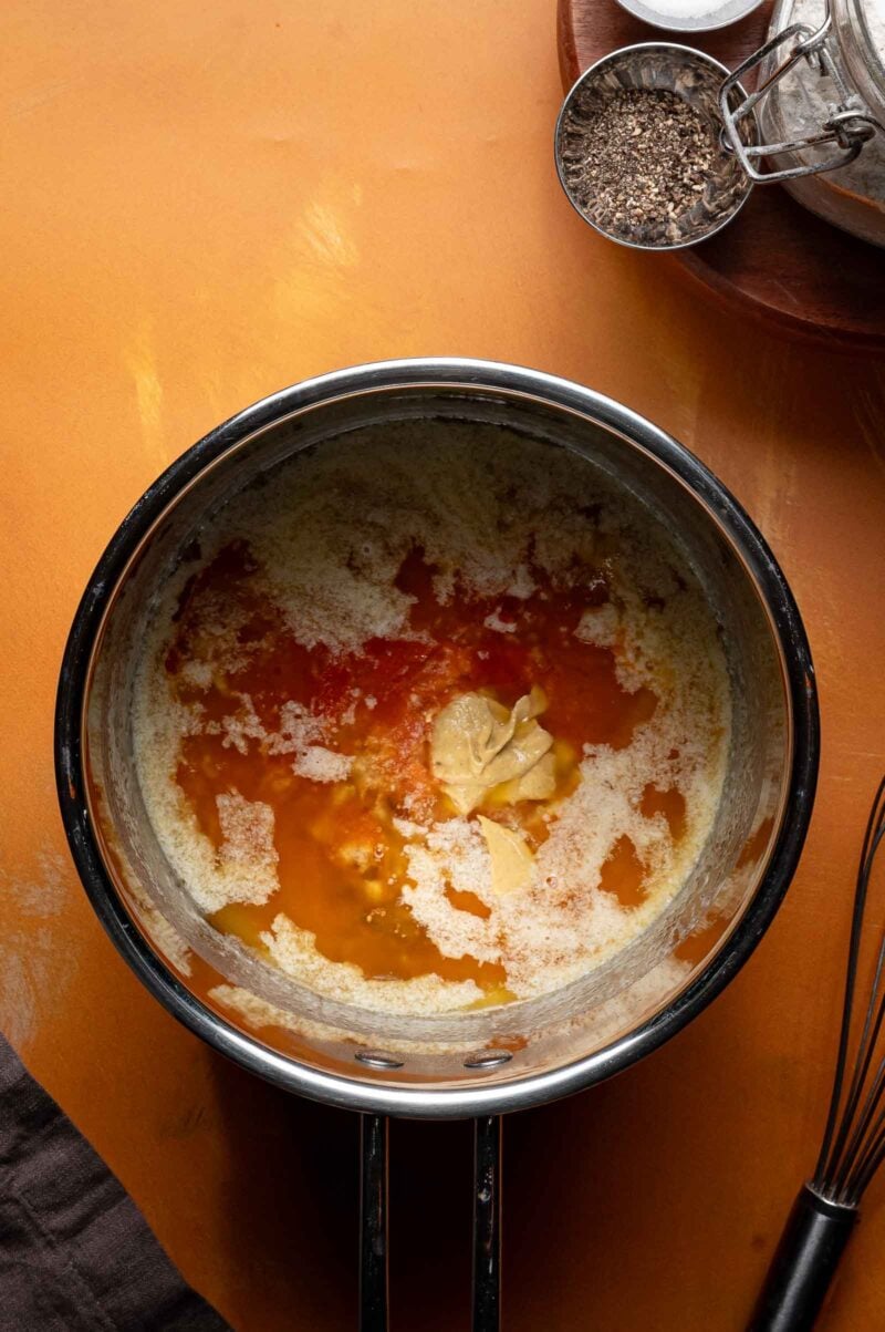 A saucepan containing melted butter, hot sauce, and mustard sits on an orange countertop next to a small bowl of black pepper and a metal whisk.