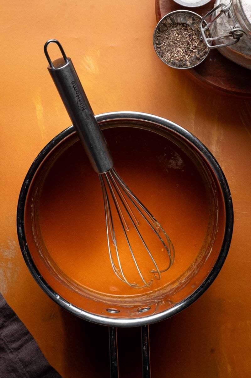 A metal whisk rests in a saucepan filled with orange buffalo sauce on an orange surface, with a tin of pepper and flour nearby.