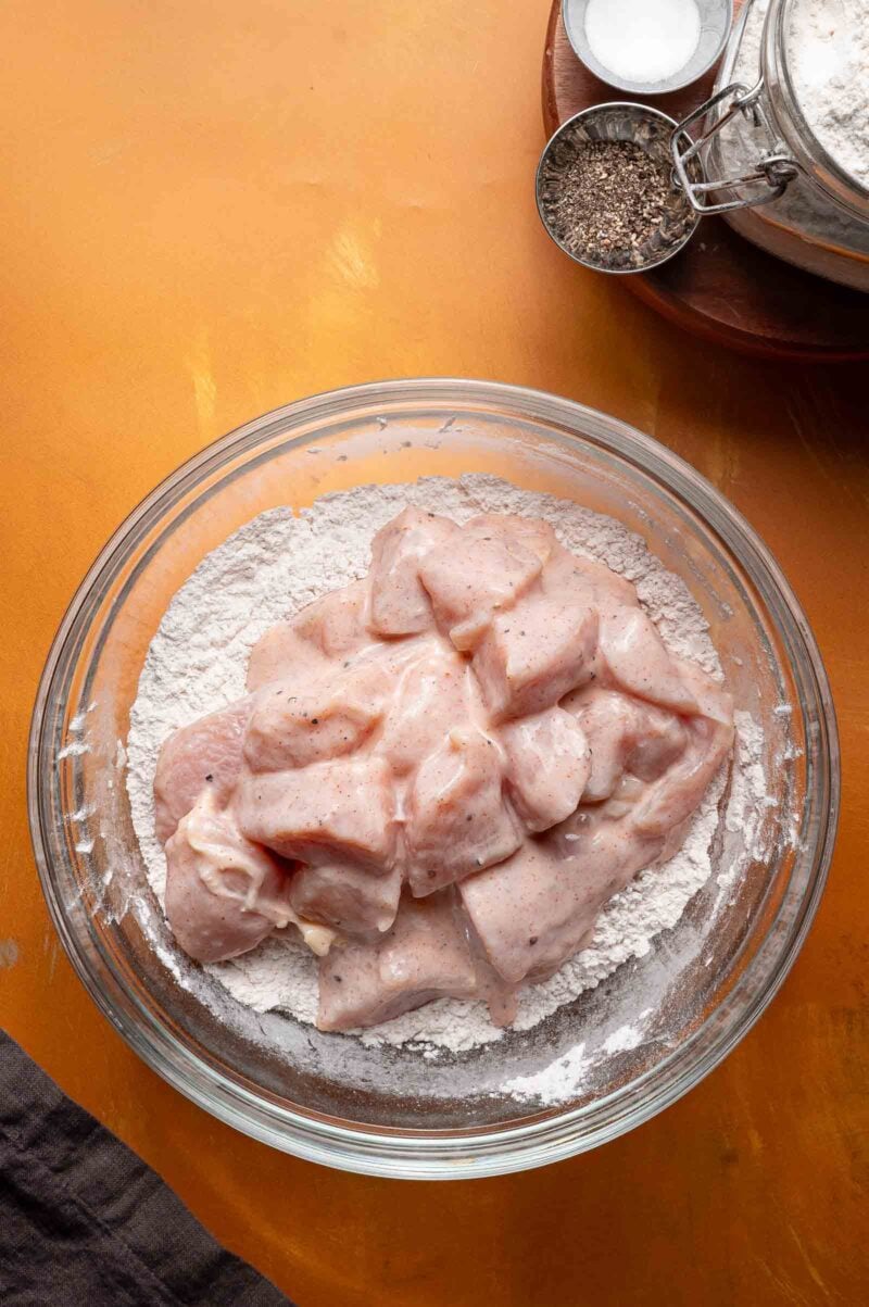 A glass bowl filled with flour and raw, seasoned chicken pieces on a yellow surface, with salt, pepper, and flour visible in the background.