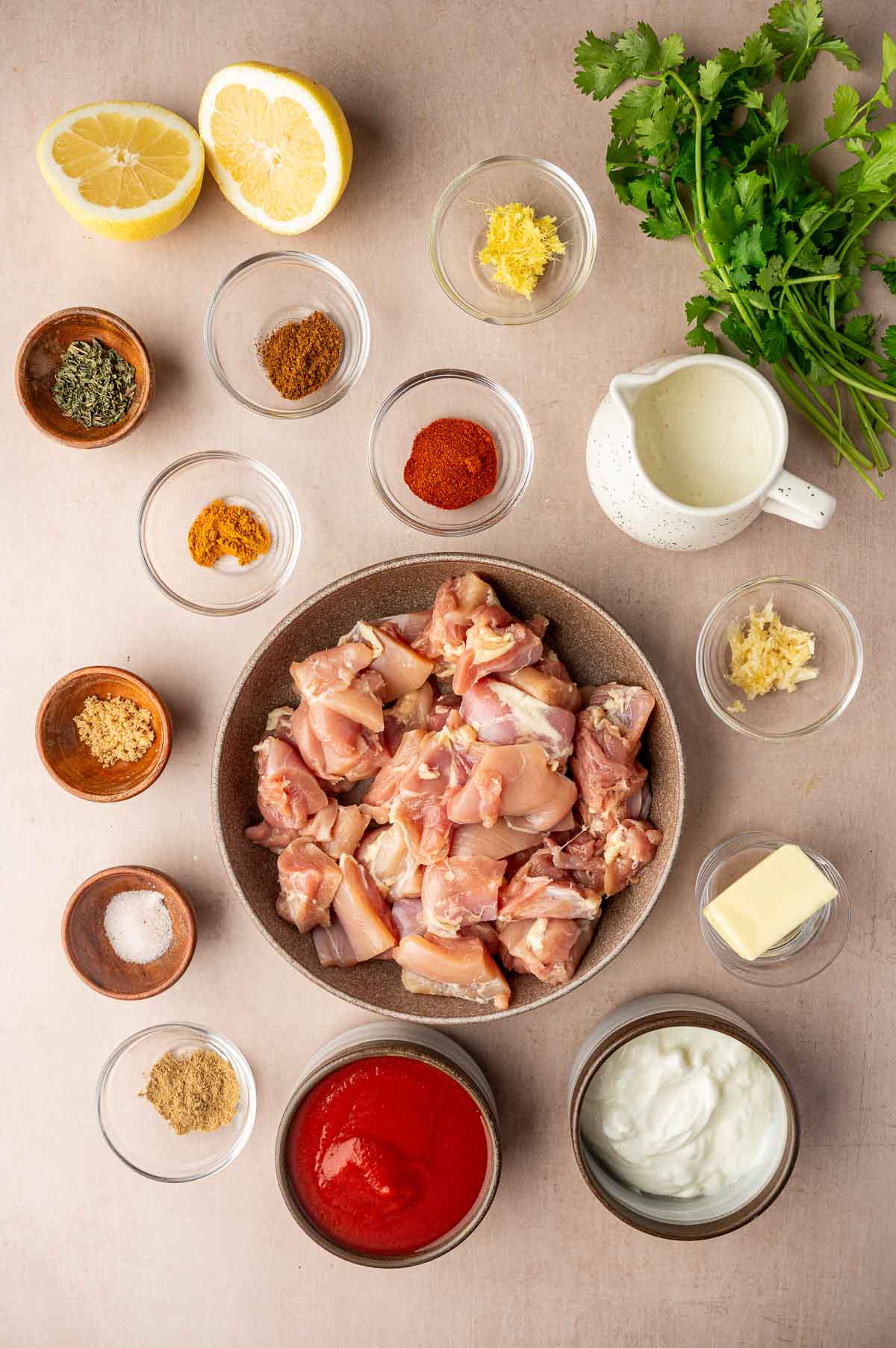 Overhead view of ingredients for butter chicken with raw chicken pieces in a bowl surrounded by various spices, herbs, lemon halves, cilantro, tomato sauce, yogurt, cream, ginger, garlic, and butter.