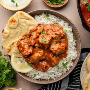 A bowl of rice topped with butter chicken, garnished with cilantro, served with naan, lemon wedges, yogurt sauce, and extra cilantro on the side.