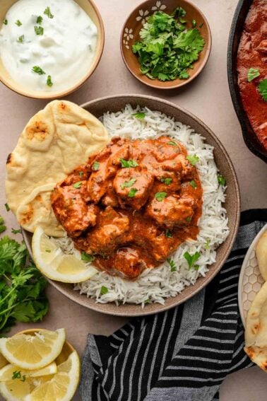 A bowl of rice topped with butter chicken, garnished with cilantro, served with naan, lemon wedges, yogurt sauce, and extra cilantro on the side.