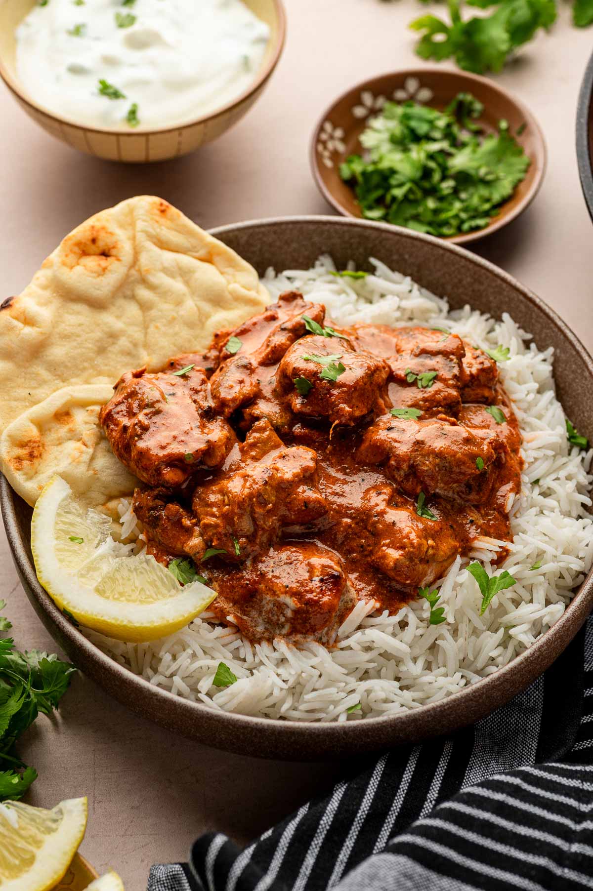 A bowl of white rice topped with pieces of butter chicken in a creamy sauce, served with naan bread, lemon wedges, and garnished with chopped herbs.