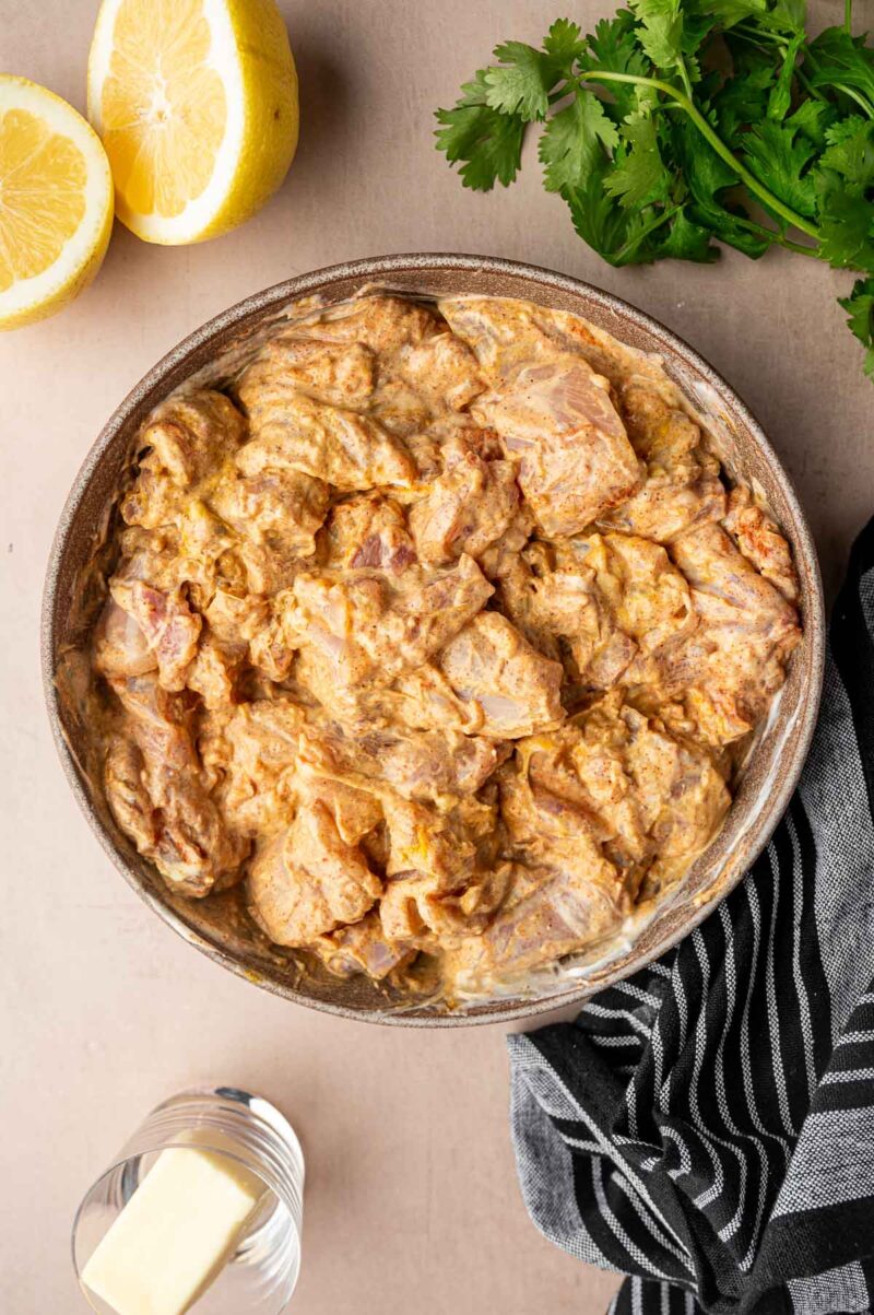 A bowl of marinated raw chicken pieces sits on a table, surrounded by lemon halves, fresh cilantro, a glass with a butter pat, and a striped cloth napkin.