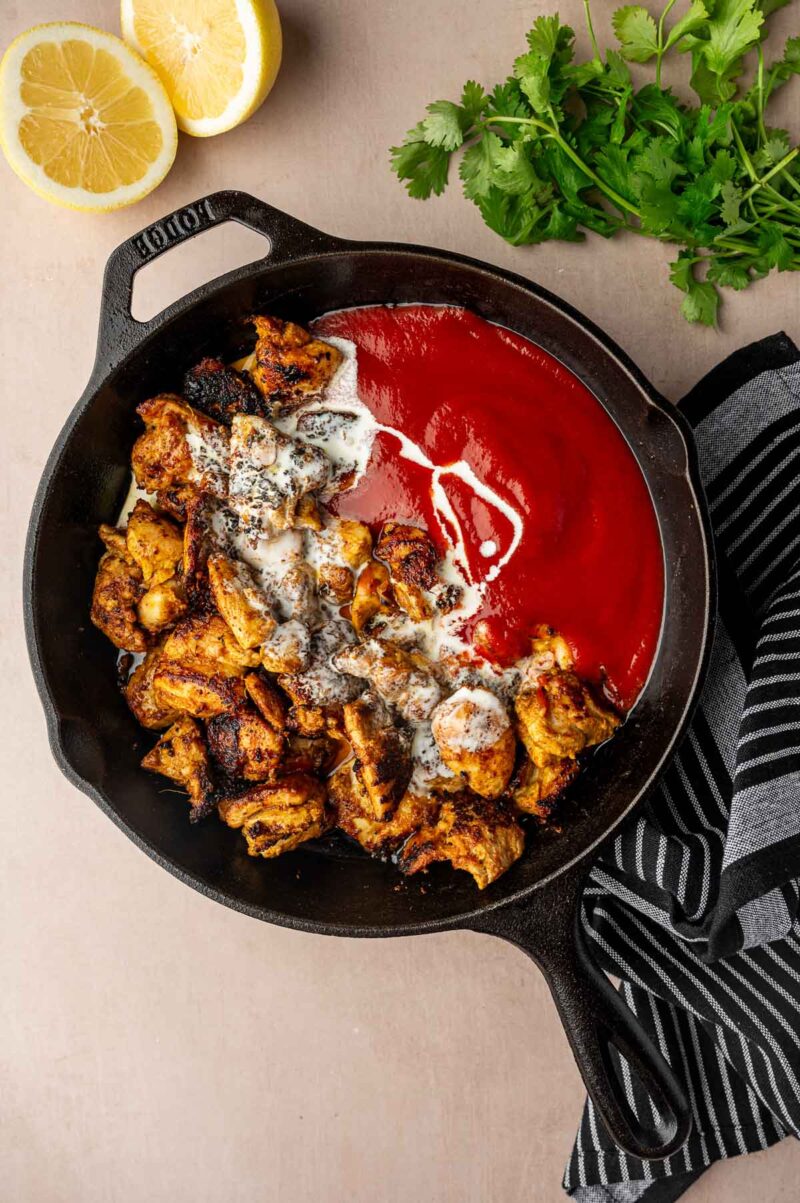 Cast iron skillet with cooked chicken pieces, red tomato puree, and cream, next to lemon halves, cilantro, and a striped towel.