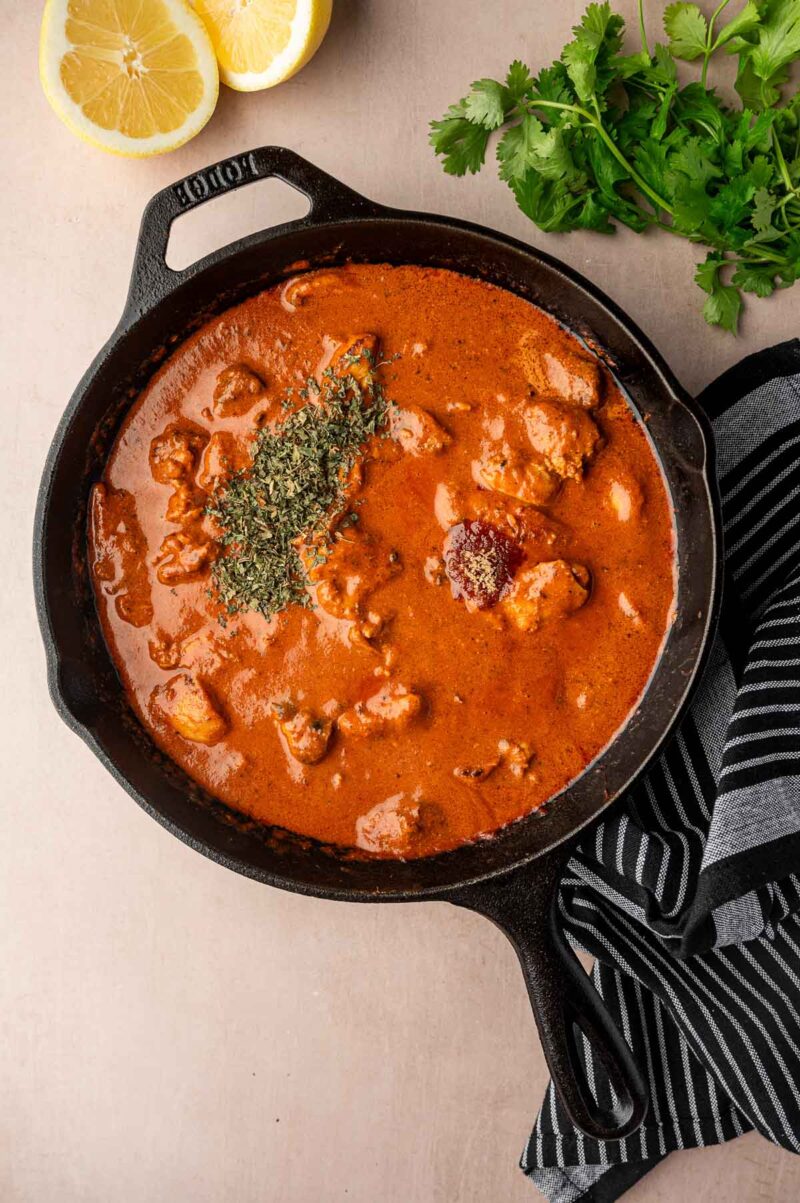 A cast iron skillet filled with creamy butter chicken with chicken pieces, topped with Kasoori methi, next to lemon halves, cilantro, and a striped cloth.