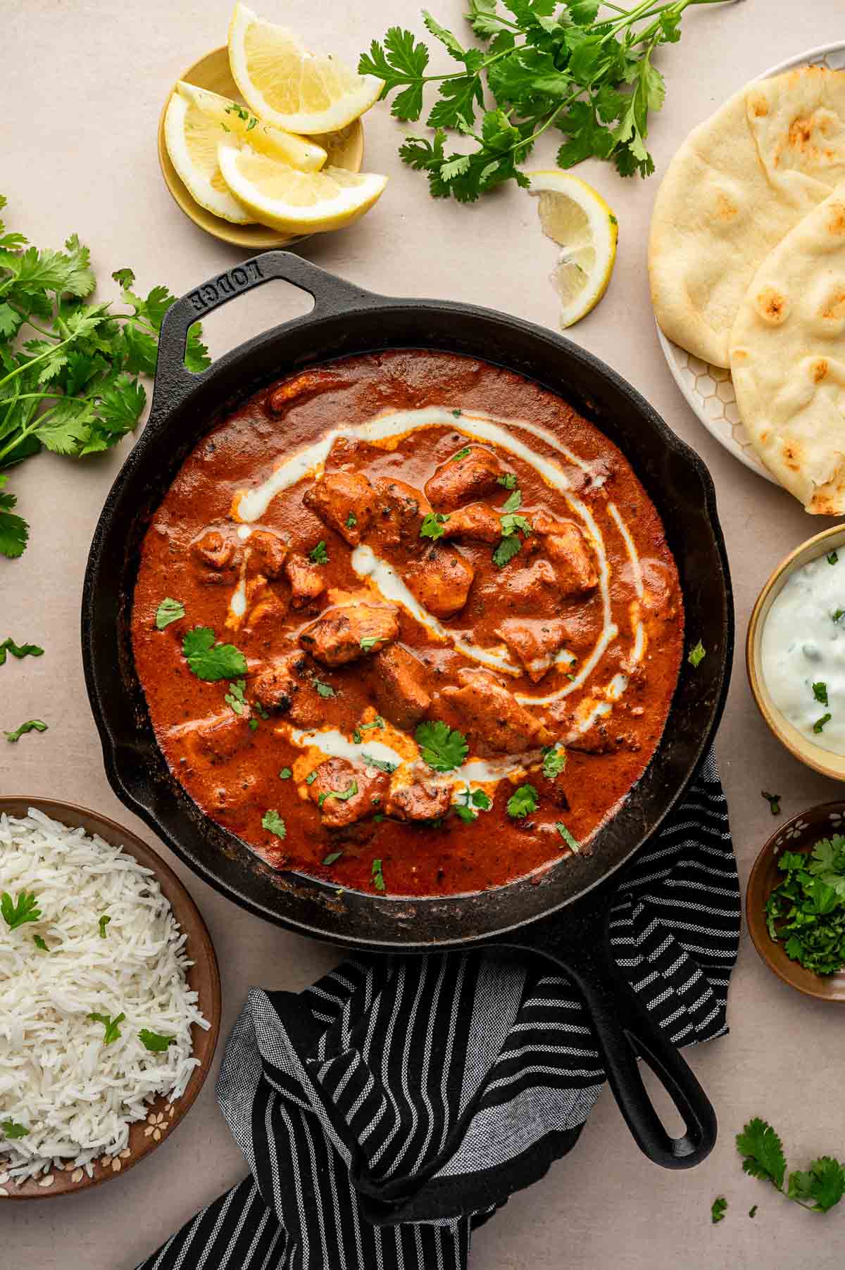 A skillet of butter chicken garnished with cream and cilantro, surrounded by naan, white rice, lemon slices, cilantro, and a bowl of yogurt sauce.