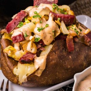 A stuffed baked potato topped with sliced corned beef, sautéed cabbage, reuben dressing, and chopped parsley, served on a white plate.