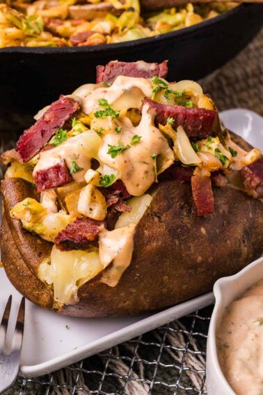 A stuffed baked potato topped with sliced corned beef, sautéed cabbage, reuben dressing, and chopped parsley, served on a white plate.
