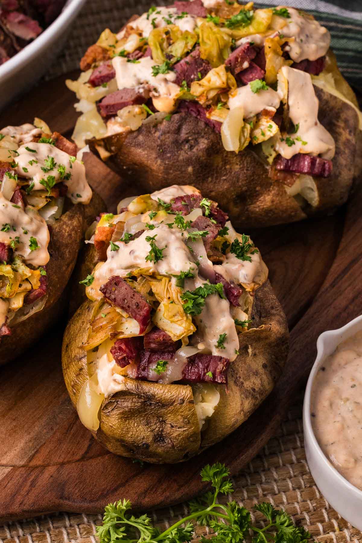 Three stuffed baked potatoes topped with chopped corned beef, fried cabbage, creamy reuben dressing, and chopped parsley are arranged on a wooden serving board.