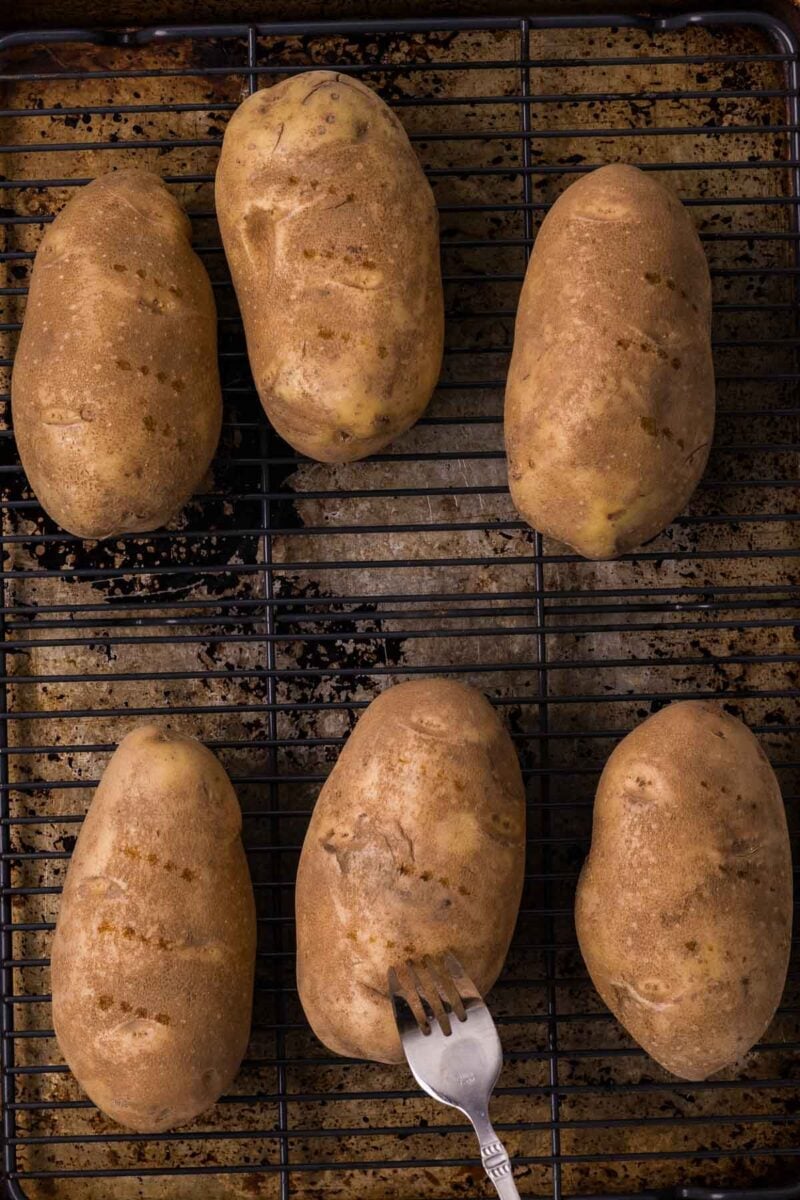 Six whole russet potatoes are placed on a wire rack over a baking sheet, with a fork piercing one potato.