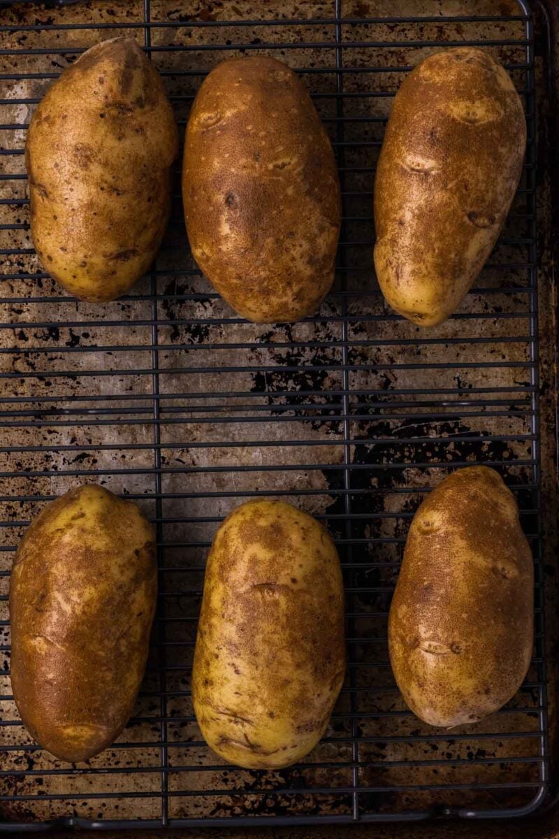 Six whole russet potatoes are arranged on a metal cooling rack placed over a baking sheet.