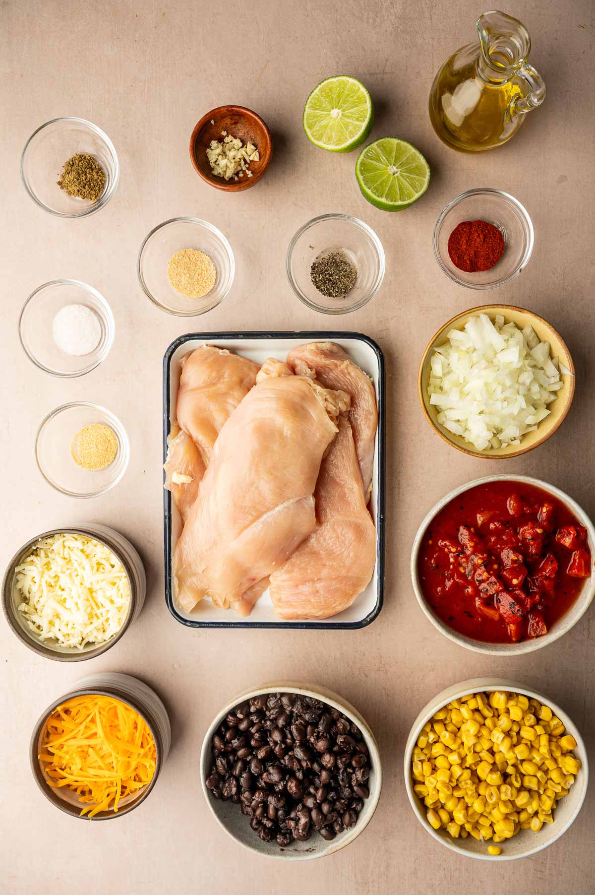 Raw chicken breasts surrounded by bowls of black beans, corn, canned tomatoes, chopped onion, shredded cheeses, spices, minced garlic, lime halves, and olive oil on a light surface.