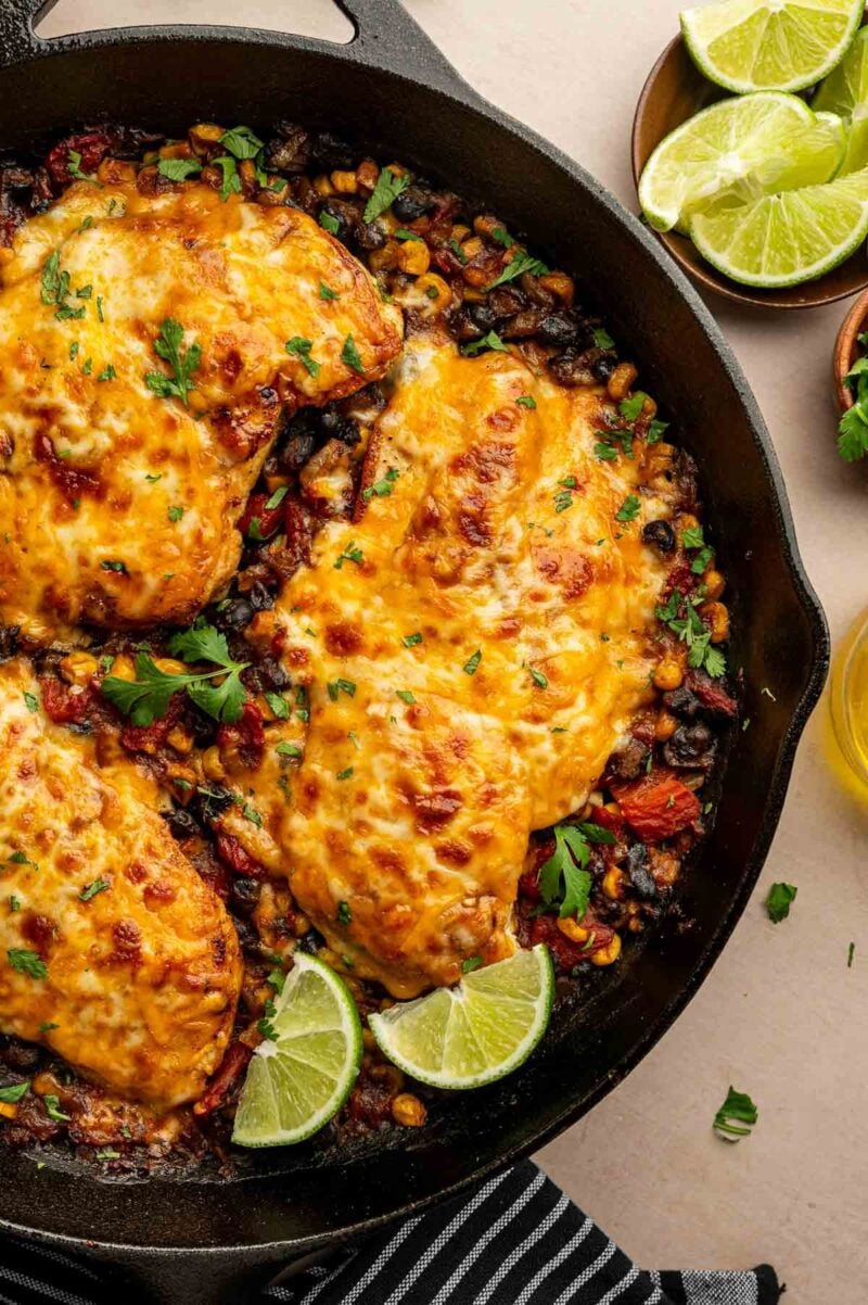 A skillet with cowboy chicken breasts topped with melted cheese, set on a bed of corn, black beans, and tomatoes, garnished with lime wedges and fresh herbs.