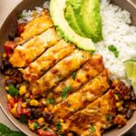 A bowl of sliced cowboy chicken breast over rice, served with black beans, corn, diced tomatoes, and avocado slices, garnished with cilantro and a lime wedge.