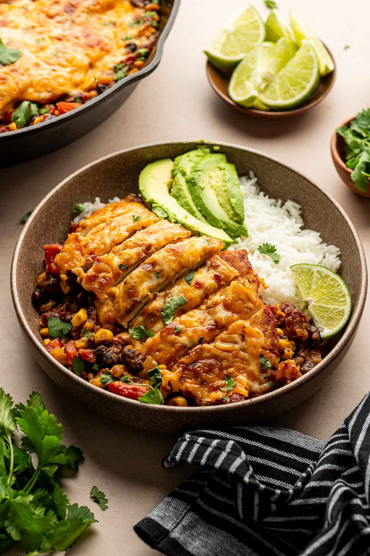 A bowl of white rice topped with cheesy cowboy chicken, black beans, corn, avocado slices, and a lime wedge. A skillet, sliced limes, and cilantro are also visible.