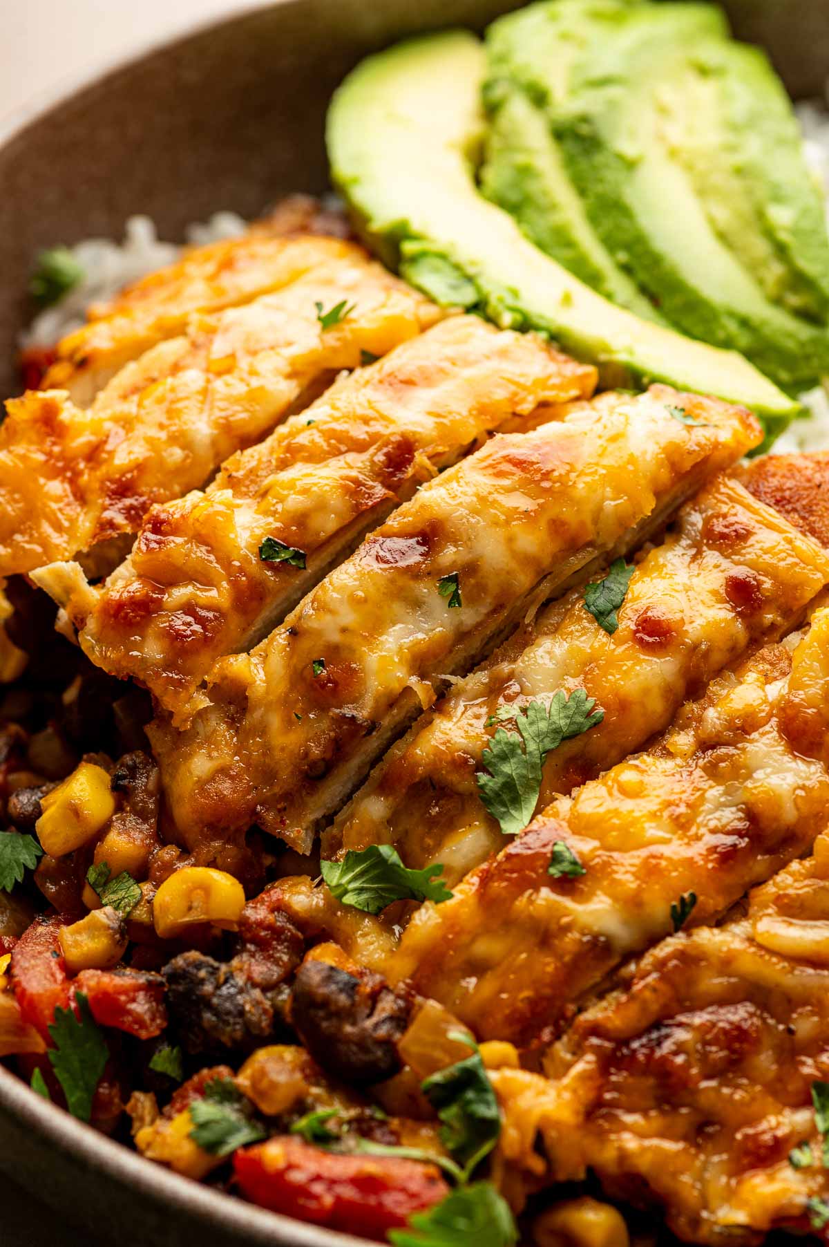 Sliced cheesy cowboy chicken breast on a bed of rice with black beans, corn, tomatoes, and avocado slices, garnished with chopped cilantro.