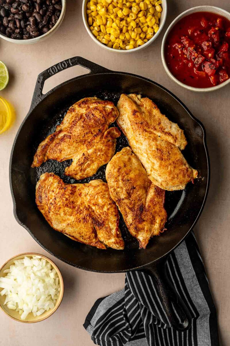 Four seasoned chicken breasts are cooking in a cast iron skillet, surrounded by bowls of black beans, corn, salsa, and chopped onions.