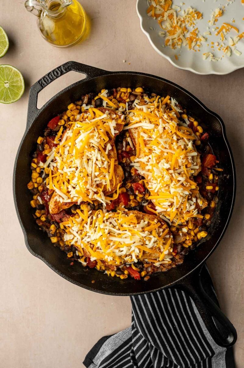 A cast iron skillet filled with a corn, black bean, and tomato mixture topped with shredded cheese. A striped towel, limes, olive oil, and a plate with extra cheese are nearby.