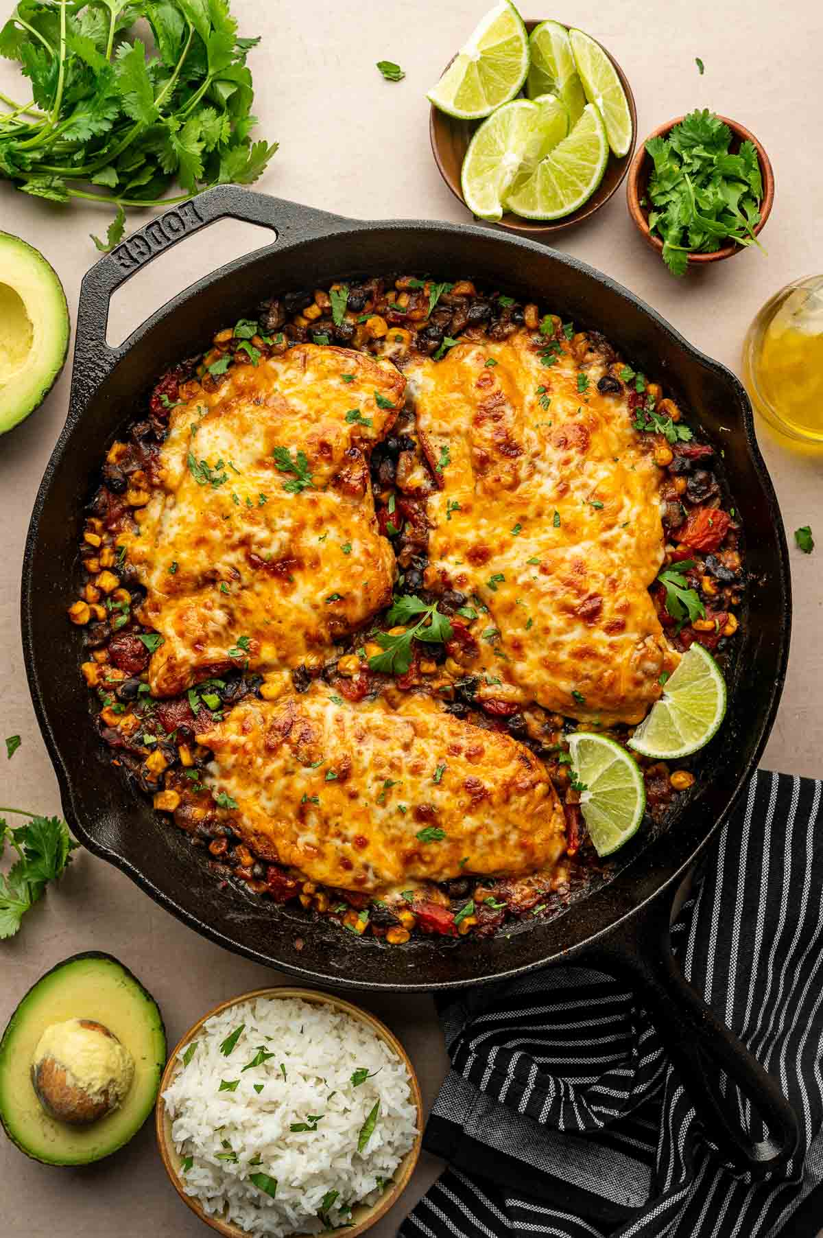A cast iron skillet with baked chicken breasts topped with melted cheese, surrounded by corn and beans, garnished with lime wedges, herbs, and served with rice and avocado.