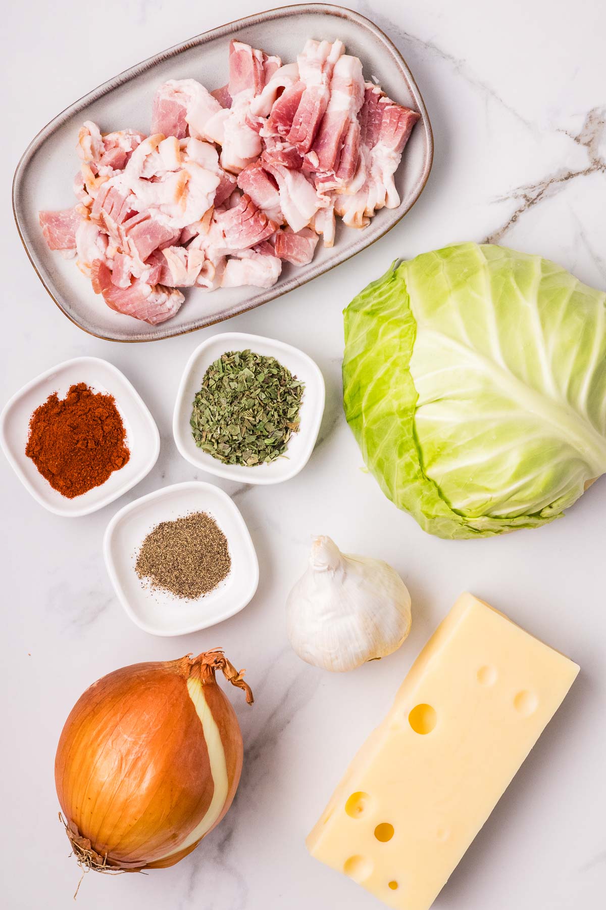 Raw bacon, cabbage, onion, garlic, Swiss cheese, dried herbs, ground pepper, and paprika arranged on a white marble surface.