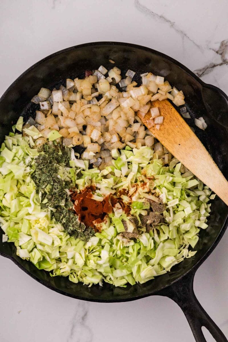 Chopped onions, cabbage, herbs, and spices are being cooked in a black cast iron skillet with a wooden spatula on a marble countertop.