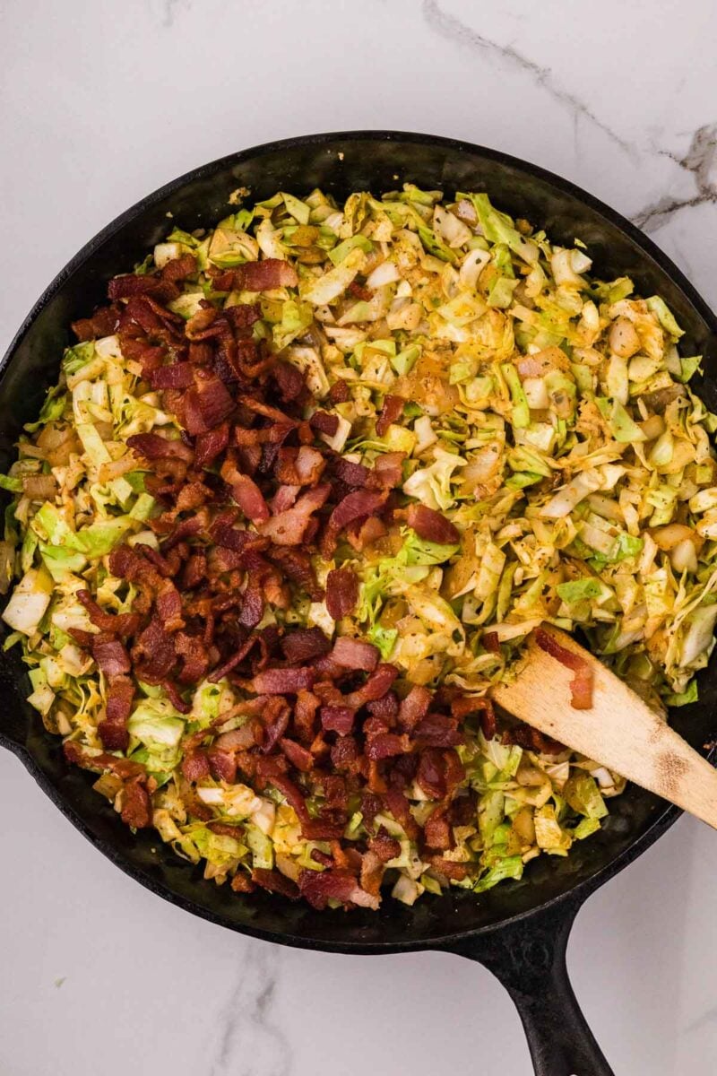 A skillet filled with fried cabbage and crispy bacon pieces, with a wooden spatula resting inside.