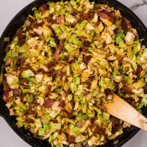 Fried cabbage and bacon cooked in a cast iron skillet, stirred with a wooden spatula, on a white marble surface.