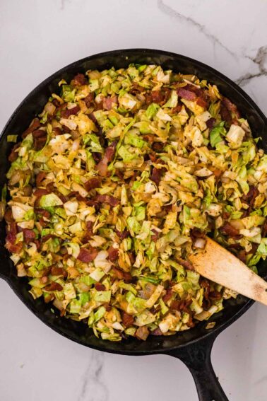 Fried cabbage and bacon cooked in a cast iron skillet, stirred with a wooden spatula, on a white marble surface.