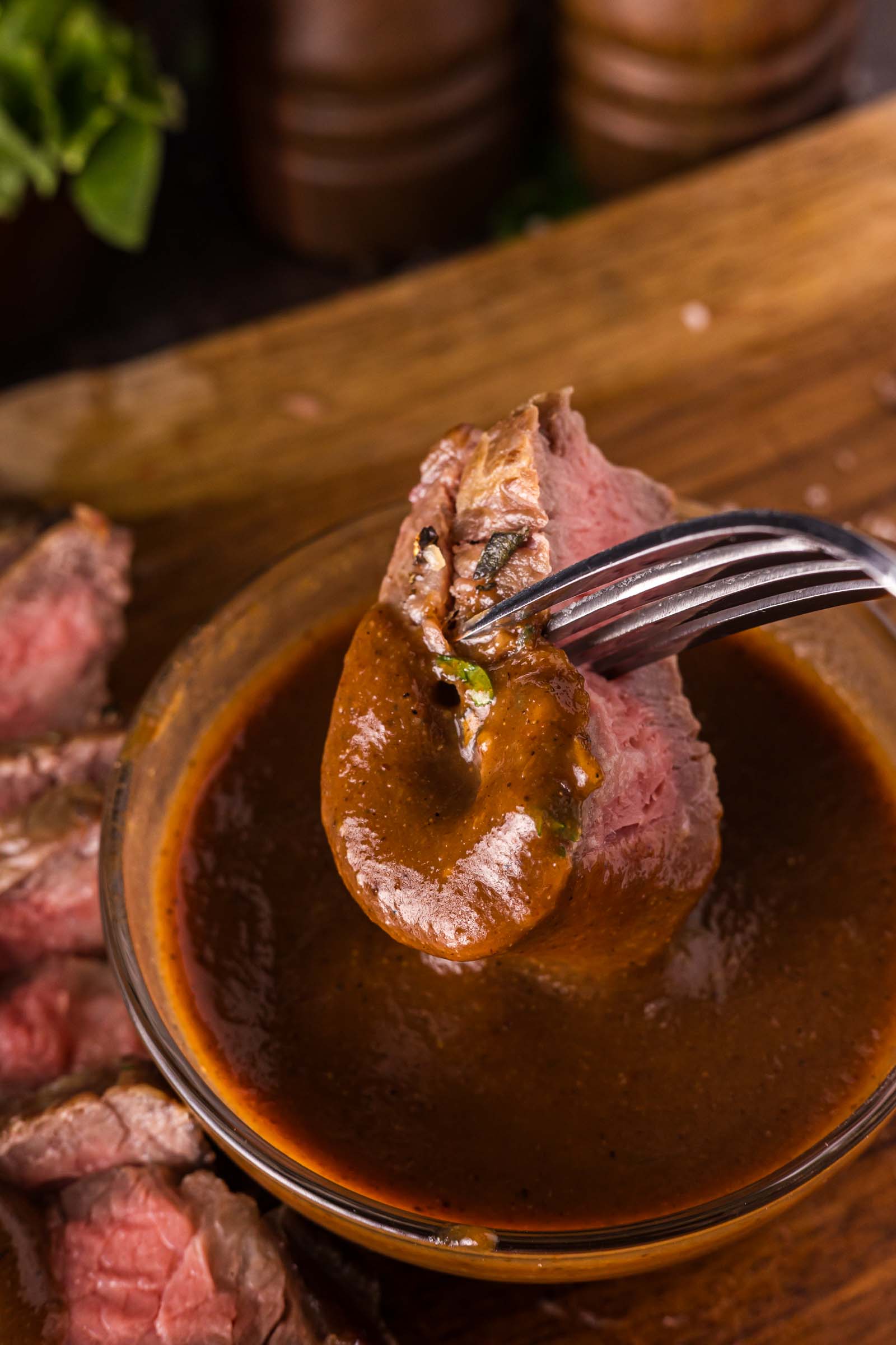 A fork holds a piece of medium-rare steak dipped in steak sauce, with more sliced steak and a wooden surface in the background.