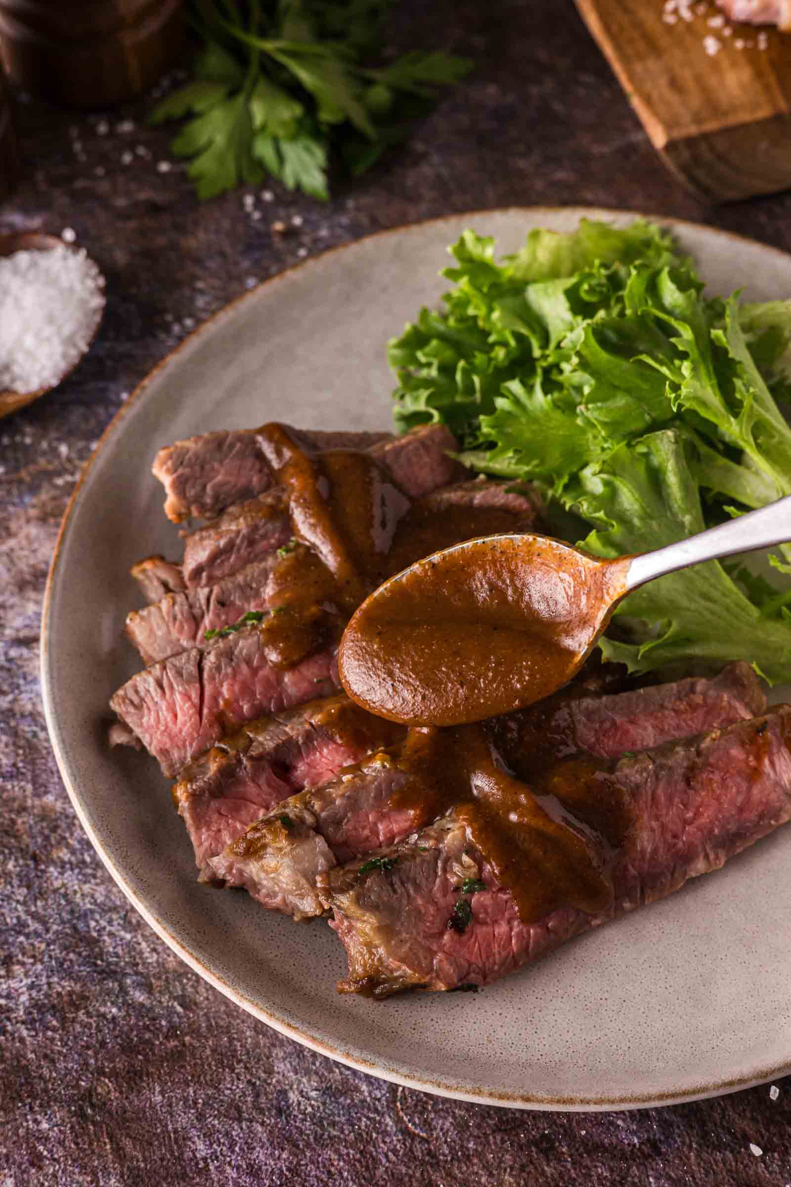 Sliced medium-rare steak on a plate with leafy greens, being topped with thick steak sauce from a spoon.