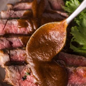 Sliced medium-rare steak on a plate with green lettuce, topped with brown homemade steak sauce being spooned over the meat.