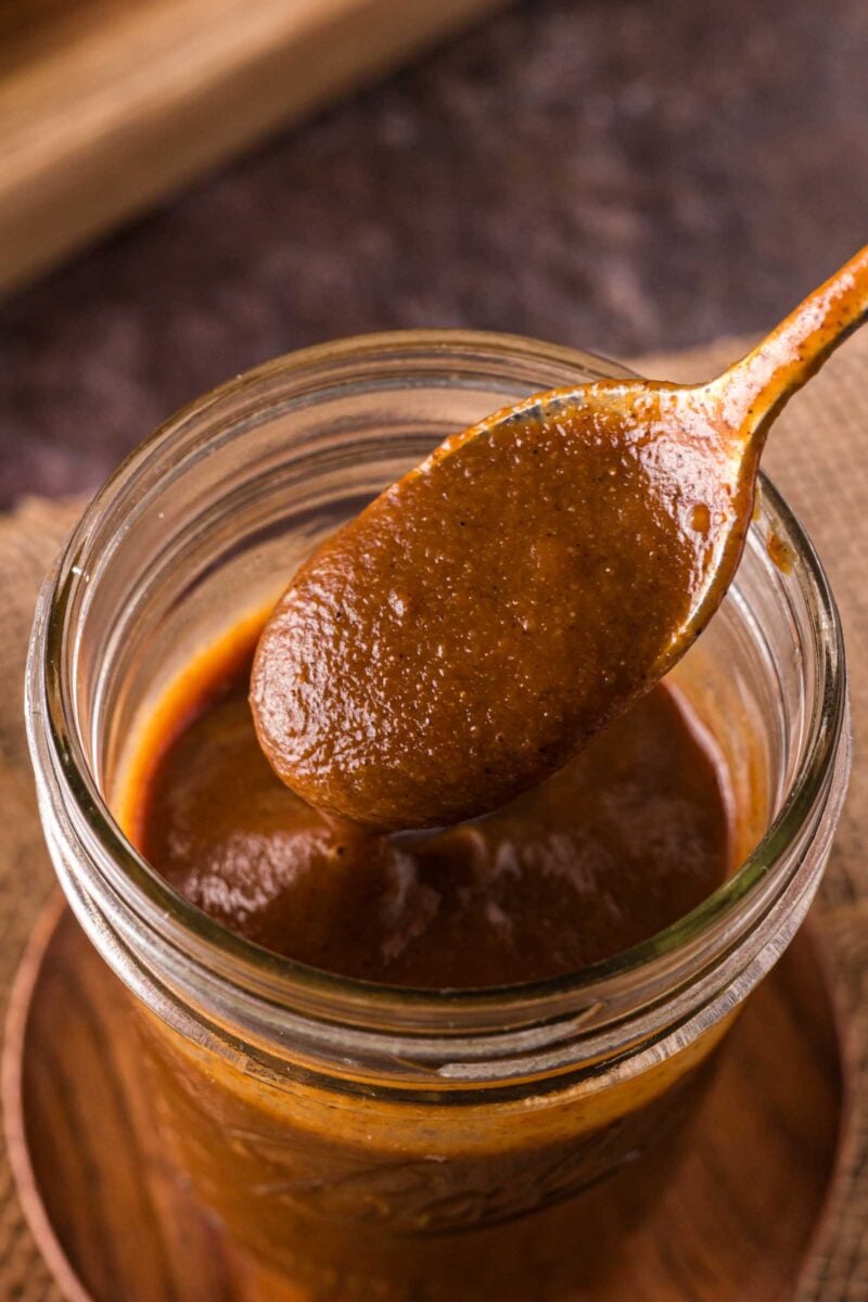 A wooden spoon holding thick a1 steak sauce above a glass jar filled with more sauce, placed on a brown surface.