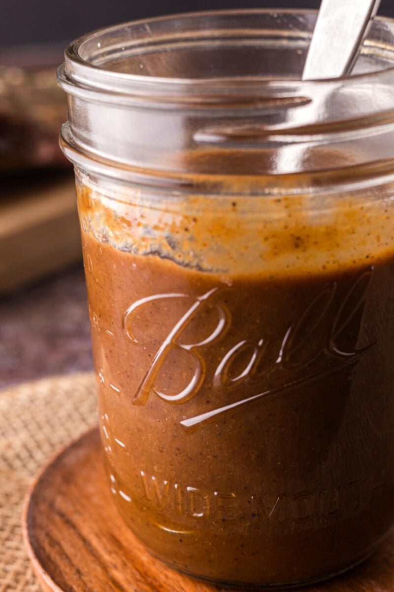 A glass Ball jar filled with brown homemade steak sauce, with a metal spoon inside, placed on a wooden coaster.