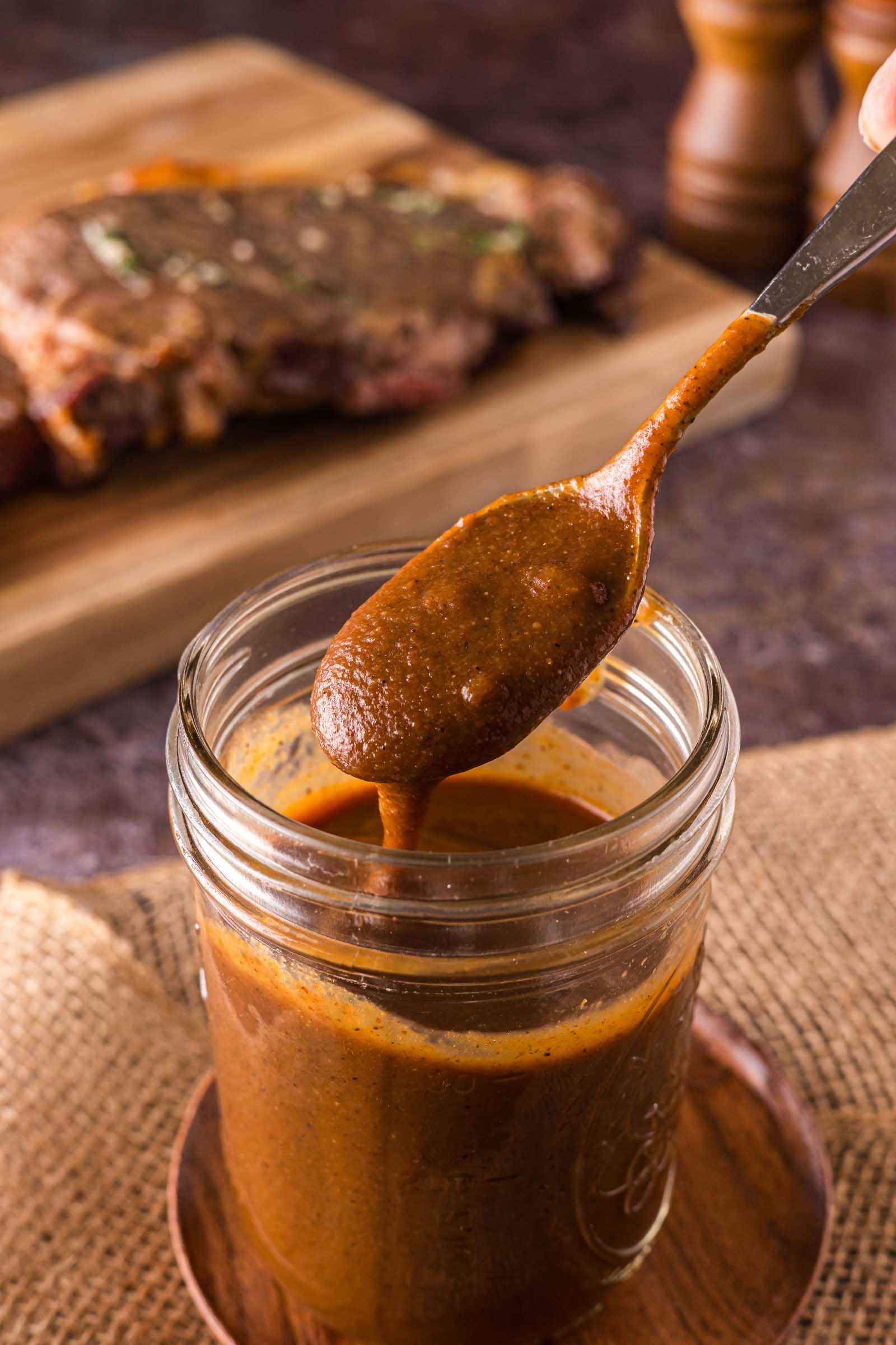 A spoon dips into a jar of thick a1 steak sauce, with a piece of cooked steak on a wooden board in the background.
